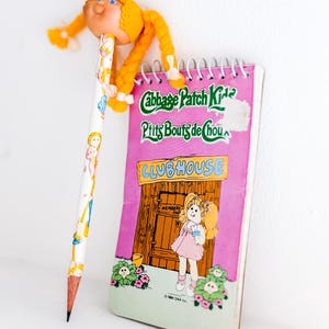 May include: A pencil with a Cabbage Patch Kids doll topper and a spiral-bound notebook. The pencil is white with illustrations of Cabbage Patch Kids. The notebook is pink with the words "Cabbage Patch Kids" and "Clubhouse" on the cover.