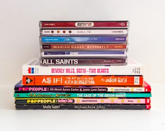 May include: A stack of CDs and books. The CDs include music by Destiny's Child, Aqua, Mariah Carey, and All Saints. The books include "Beverly Hills, 90210 - Two Hearts", "As If!", "PopPeople: All About Aaron Carter & Jamie Lynn Spears", "The Case of the Green Ghost", and "Totally Taylor!"