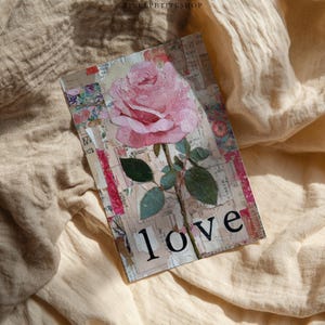 May include: A rectangular card featuring a pink rose with green leaves and the word "love" in black. The card has a collage-like background with floral and text elements. The card is set against a beige, textured fabric.