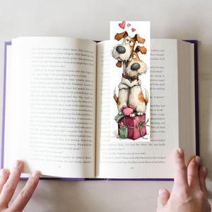 May include: A whimsical bookmark featuring two cartoon dogs embracing, with hearts floating above them. The dogs are brown and white, sitting atop a red gift box. The bookmark is placed in an open book with purple edges.