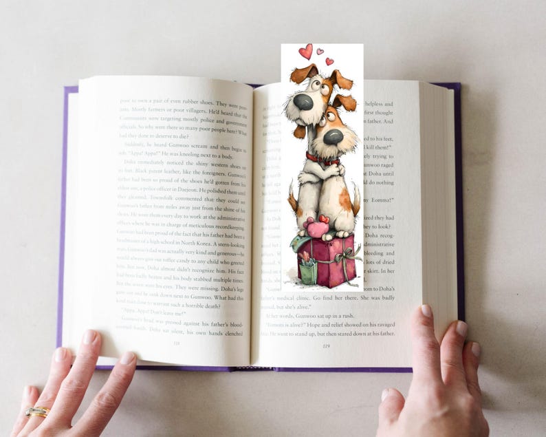 May include: A whimsical bookmark featuring two cartoon dogs embracing, with hearts floating above them. The dogs are brown and white, sitting atop a red gift box. The bookmark is placed in an open book with purple edges.