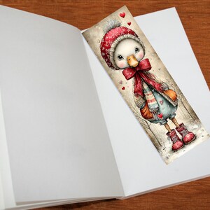 May include: A whimsical bookmark featuring a cartoon duck wearing a red winter hat, scarf, and boots. The duck is standing in a snowy scene with hearts. The bookmark is placed inside an open book.
