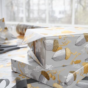May include: Two wrapped gifts with gray paper featuring gold and silver acorn and leaf designs. A pair of scissors and a roll of matching wrapping paper are visible, suggesting a gift-wrapping theme.