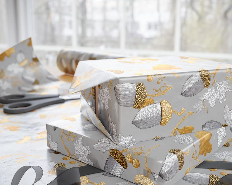May include: Two wrapped gifts with gray paper featuring gold and silver acorn and leaf designs. A pair of scissors and a roll of matching wrapping paper are visible, suggesting a gift-wrapping theme.