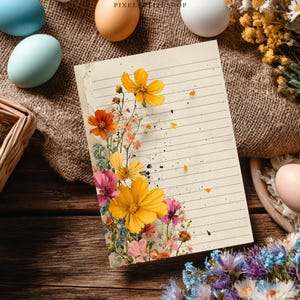 May include: A lined paper with a floral design featuring yellow, orange, and pink flowers. The paper is surrounded by Easter eggs in various colors, a woven basket, and dried flowers, all arranged on a wooden surface.