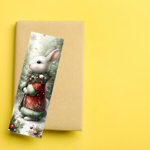 Puede incluir: Un marcap&aacute;ginas navide&ntilde;o con un conejo blanco con un abrigo rojo y ribete de piel verde, sosteniendo un bast&oacute;n. El conejo lleva una corona festiva. El marcap&aacute;ginas est&aacute; sobre un libro marr&oacute;n, sobre un fondo amarillo.