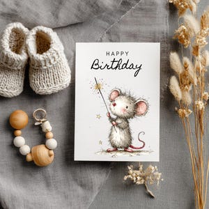 May include: A greeting card with a watercolor illustration of a mouse holding a sparkler, with the words "Happy Birthday". Beside it are a pair of knitted baby booties and a wooden teething ring. Dried flowers are also present.