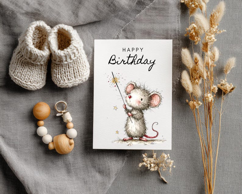 May include: A greeting card with a watercolor illustration of a mouse holding a sparkler, with the words "Happy Birthday". Beside it are a pair of knitted baby booties and a wooden teething ring. Dried flowers are also present.