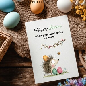 Pode incluir: Um cart&atilde;o de P&aacute;scoa com uma ilustra&ccedil;&atilde;o de ouri&ccedil;o, ovos coloridos e o texto "Happy Easter". O cart&atilde;o &eacute; rodeado por ovos decorativos e flores numa superf&iacute;cie de madeira.