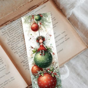 Puede incluir: Marcap&aacute;ginas con tem&aacute;tica navide&ntilde;a con una ilustraci&oacute;n en acuarela. Un hada con un vestido rojo est&aacute; sentada sobre una bola verde, rodeada de adornos rojos y verdes. El marcap&aacute;ginas est&aacute; sobre un libro abierto.