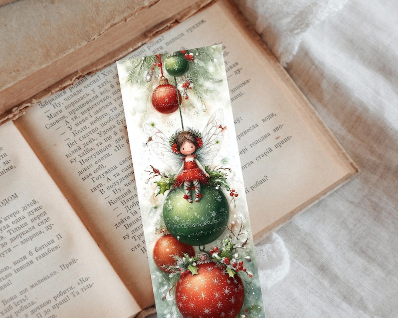 Puede incluir: Marcap&aacute;ginas con tem&aacute;tica navide&ntilde;a con una ilustraci&oacute;n en acuarela. Un hada con un vestido rojo est&aacute; sentada sobre una bola verde, rodeada de adornos rojos y verdes. El marcap&aacute;ginas est&aacute; sobre un libro abierto.