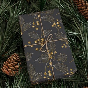 May include: A wrapped gift with a dark gray background and a gold holly and berry pattern. The gift is tied with twine and rests on a bed of green pine needles and pine cones, creating a festive holiday scene.