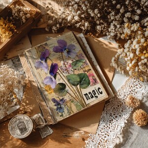 May include: A vintage-style journal with pressed purple violets and green leaves on the cover, with the word "magic" printed on the front. The journal is surrounded by dried flowers, lace, and antique ephemera.