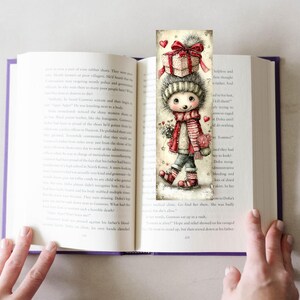 May include: A whimsical bookmark featuring a cartoon character with a gift box on its head, wearing a striped scarf and holding flowers. The bookmark is inserted into an open book, with the pages visible.