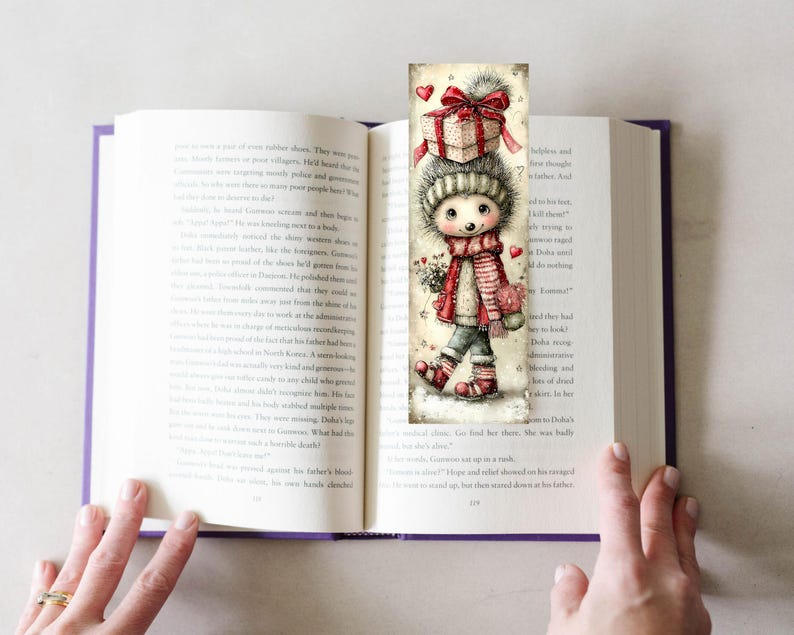 May include: A whimsical bookmark featuring a cartoon character with a gift box on its head, wearing a striped scarf and holding flowers. The bookmark is inserted into an open book, with the pages visible.