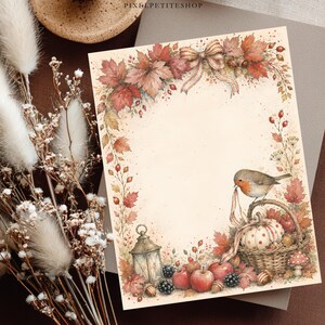 Op de afbeelding: Een rechthoekig vel briefpapier met een aquarelillustratie van herfstbladeren, rode bessen, een roodborstje op een mand met een pompoen met stippen, een lantaarn en appels, omlijst door een decoratieve strik aan de bovenkant in het midden.