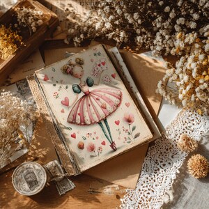 Może przedstawiać: Dziennik w stylu vintage z ilustracją baletnicy na okładce. Baletnica ma na sobie r&oacute;żowo-białą sukienkę, otoczona sercami i kwiatami. Otwarty dziennik ukazuje strony w linie, otoczony suszonymi kwiatami i koronką.