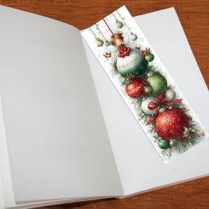 Puede incluir: Marcap&aacute;ginas con tem&aacute;tica navide&ntilde;a con una ilustraci&oacute;n en acuarela. El dise&ntilde;o incluye un hada, adornos rojos y verdes y un lazo rojo. El marcap&aacute;ginas est&aacute; colocado dentro de un libro abierto.