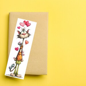 May include: A whimsical bookmark featuring a cartoon cat holding hearts and small creatures. The cat has a long neck and is wearing an orange shirt and green pants. The bookmark is placed on a brown book against a yellow background.