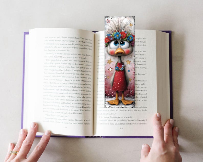 May include: A decorative bookmark with a cartoon duck wearing a floral crown and a red dress, inserted into an open book. The duck has large blue eyes and is surrounded by stars. A charming addition to any book.