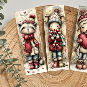 May include: Three illustrated bookmarks featuring adorable winter animals. The raccoon wears a red scarf and hat, the mouse holds a candy cane, and the lamb holds a heart. Each bookmark is set against a snowy background with hearts.