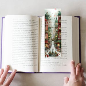 Puede incluir: Un marcap&aacute;ginas colorido con una ilustraci&oacute;n detallada en acuarela de una plaza europea con una fuente, tiendas y edificios. El marcap&aacute;ginas est&aacute; insertado en un libro abierto.