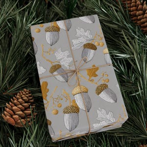 May include: A wrapped gift with a gray background featuring a pattern of acorns and leaves in silver and gold. The gift is tied with twine and rests on a bed of green pine needles and pine cones.