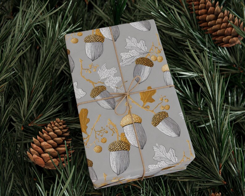 May include: A wrapped gift with a gray background featuring a pattern of acorns and leaves in silver and gold. The gift is tied with twine and rests on a bed of green pine needles and pine cones.