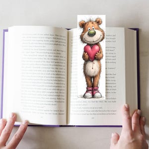 May include: A whimsical bookmark featuring a cartoon bear holding a pink heart. The bear has brown fur, a green nose, and is wearing pink sneakers. The bookmark is placed in an open book with purple edges.