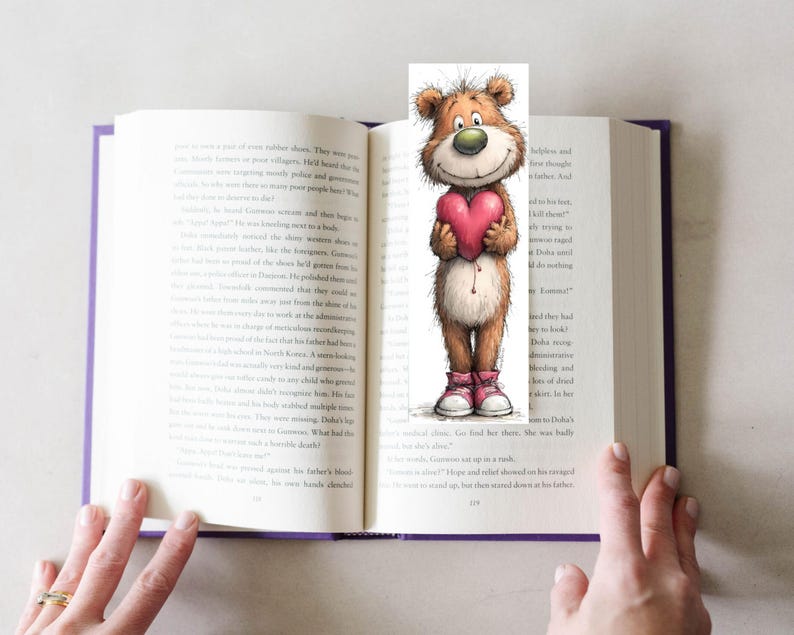 May include: A whimsical bookmark featuring a cartoon bear holding a pink heart. The bear has brown fur, a green nose, and is wearing pink sneakers. The bookmark is placed in an open book with purple edges.