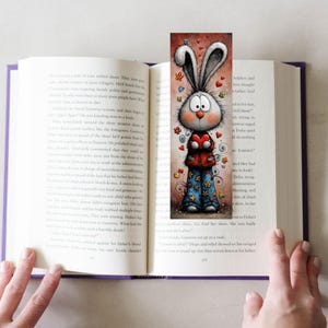 May include: A colourful bookmark with a cartoon rabbit holding a heart, placed in a book. The rabbit has big eyes, long ears, and wears a red jacket and blue trousers with star and flower patterns. Hearts and flowers are scattered around the rabbit.