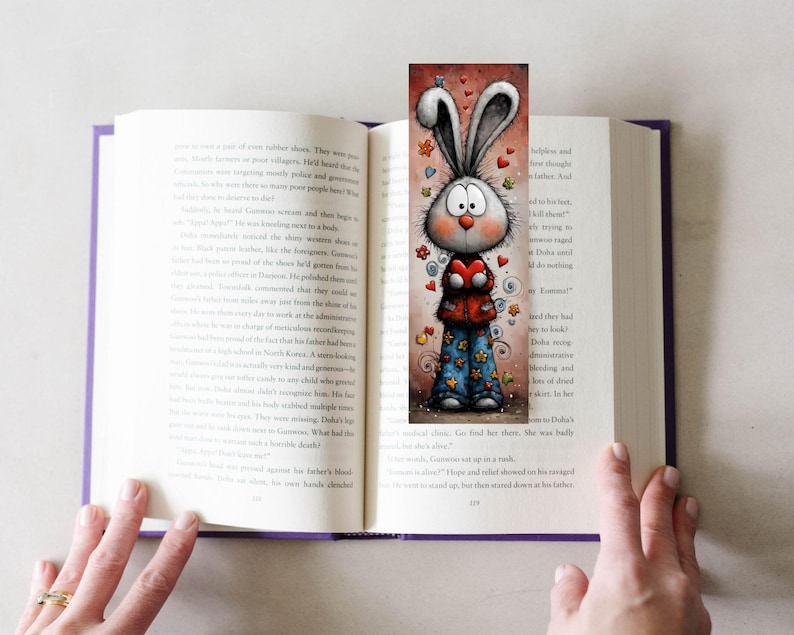 May include: A colourful bookmark with a cartoon rabbit holding a heart, placed in a book. The rabbit has big eyes, long ears, and wears a red jacket and blue trousers with star and flower patterns. Hearts and flowers are scattered around the rabbit.