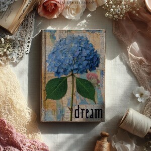 May include: A decorative wooden book with a blue hydrangea flower design. The word "dream" is written in black script. The book is surrounded by lace, flowers, and other vintage-style items.