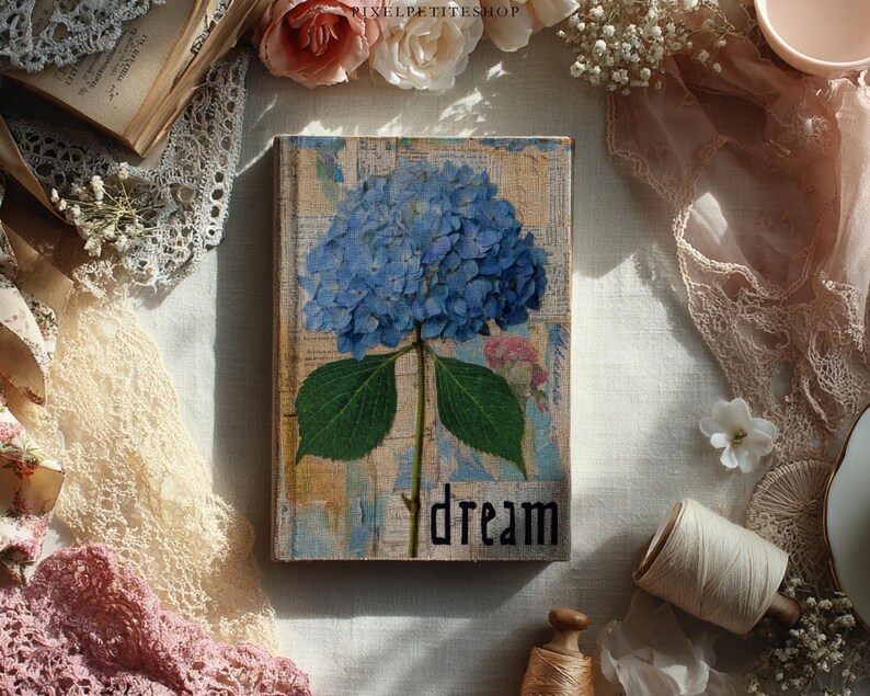 May include: A decorative wooden book with a blue hydrangea flower design. The word "dream" is written in black script. The book is surrounded by lace, flowers, and other vintage-style items.