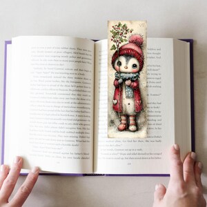May include: A whimsical bookmark featuring a cute penguin character in a winter outfit, holding a sprig of berries. The penguin wears a red coat, hat, and scarf. The bookmark is placed in an open book with purple edges.