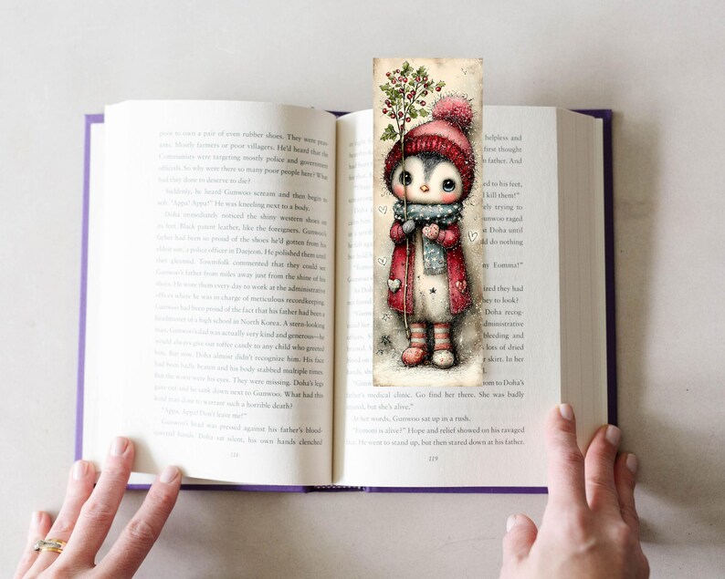 May include: A whimsical bookmark featuring a cute penguin character in a winter outfit, holding a sprig of berries. The penguin wears a red coat, hat, and scarf. The bookmark is placed in an open book with purple edges.