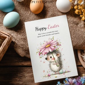 May include: An Easter greeting card with a watercolor illustration of a hedgehog holding a flower umbrella. The card reads "Happy Easter" with a message below. Pastel-colored eggs and flowers surround the card on a wooden surface.