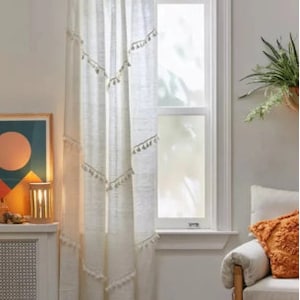 May include: Off-white linen curtain with a gold rod, hanging in front of a window. The curtain has a geometric design with tassels. The room has a rug, a chair, and a side table with a lamp.