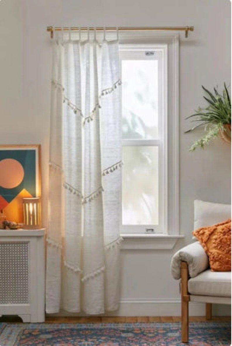 May include: Off-white linen curtain with a gold rod, hanging in front of a window. The curtain has a geometric design with tassels. The room has a rug, a chair, and a side table with a lamp.