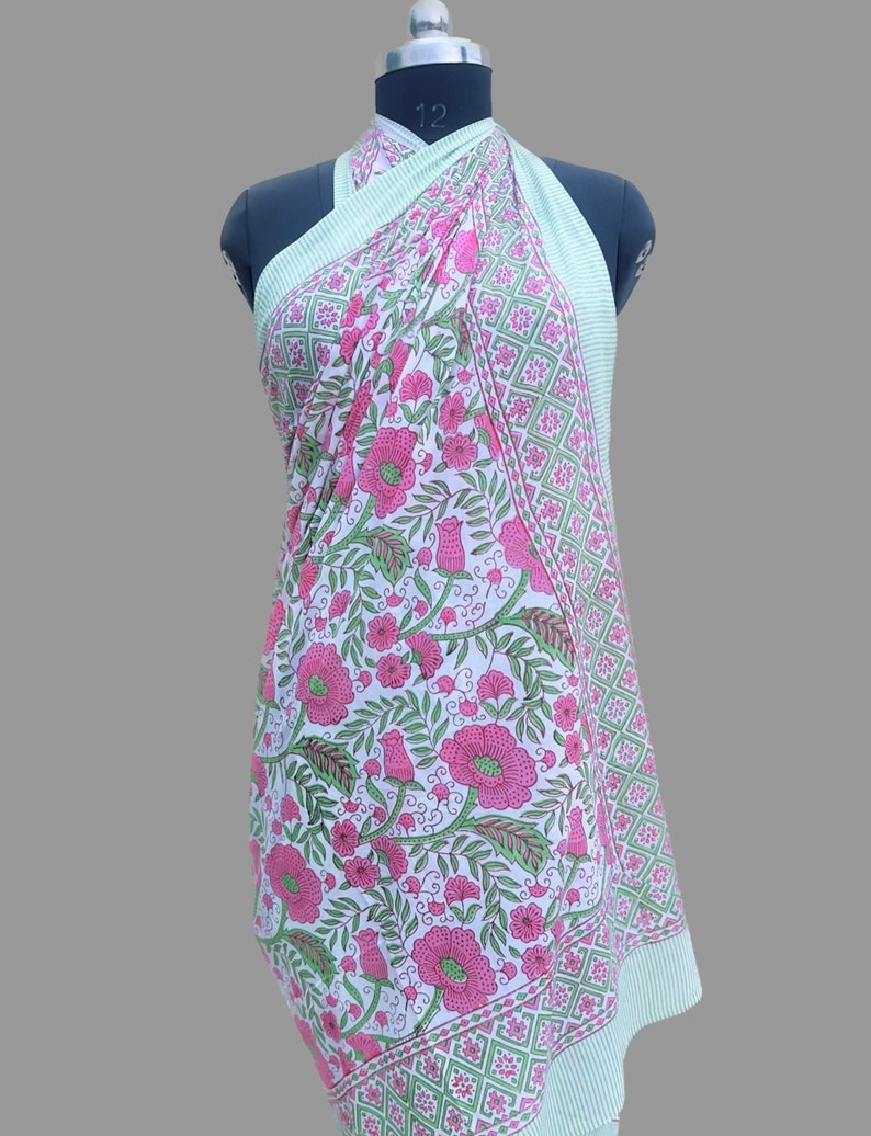 Op de afbeelding: Een groene en witte katoenen sarong met een roze bloemenprint. De sarong is om een ​​mannequin gewikkeld en heeft een rand met een ruitpatroon.
