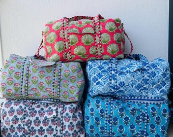 May include: A stack of five quilted duffel bags in various colors and patterns. The top bag is pink with a green shell design. Other bags feature floral and geometric patterns in blue, white, and teal.