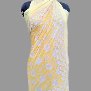 Op de afbeelding: Een gele katoenen sarong met een witte bloemenprint en een witte en gele rand. De sarong is over een mannequin gedrapeerd.