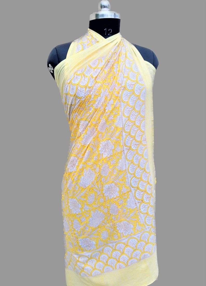 Op de afbeelding: Een gele katoenen sarong met een witte bloemenprint en een witte en gele rand. De sarong is over een mannequin gedrapeerd.