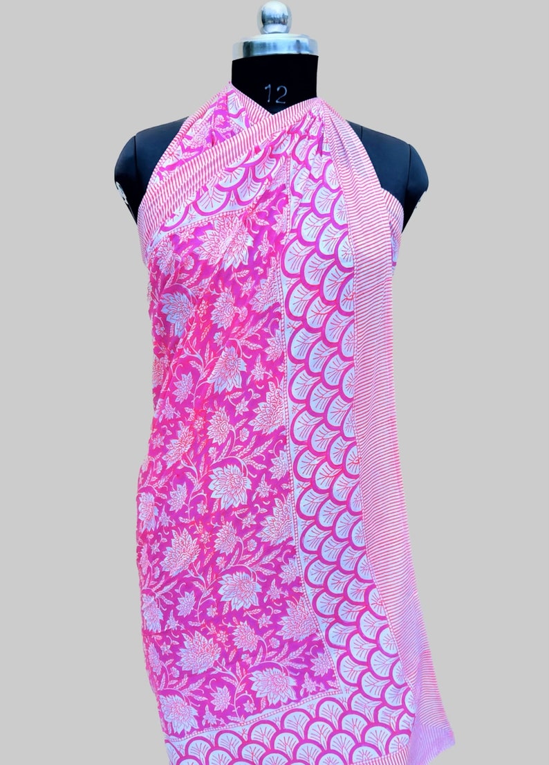 Op de afbeelding: Roze en witte katoenen sarong met een bloemenprint en een rand van witte en roze schelpen. De sarong is over een mannequin gedrapeerd.
