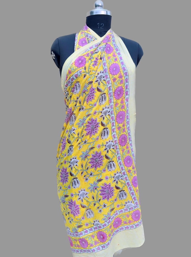 Op de afbeelding: Gele sarong met bloemenprint met roze en witte accenten. De sarong is gedrapeerd op een mannequin en is om de nek gebonden.