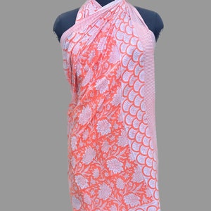 Op de afbeelding: Een perzikkleurige sarong met een witte bloemenprint en een witte rand met een gegolfd ontwerp. De sarong is over een mannequin gedrapeerd.