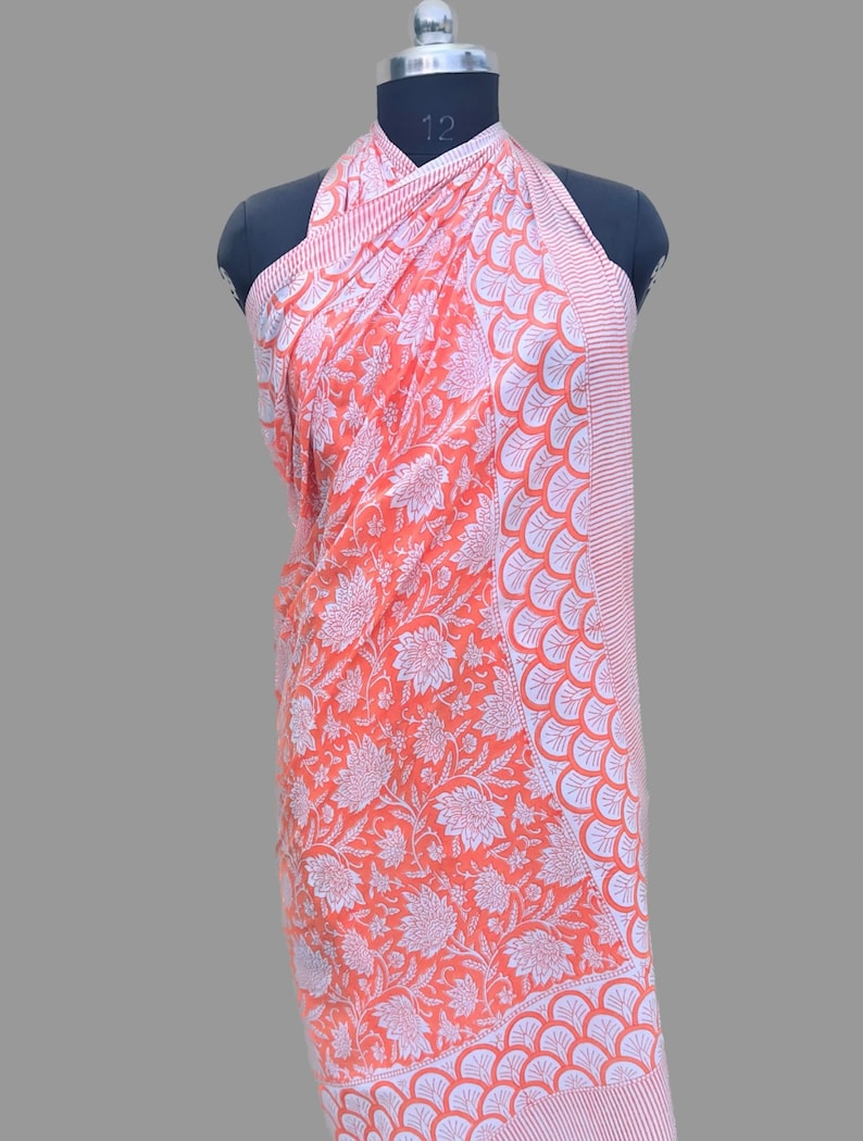 Op de afbeelding: Een perzikkleurige sarong met een witte bloemenprint en een witte rand met een gegolfd ontwerp. De sarong is over een mannequin gedrapeerd.