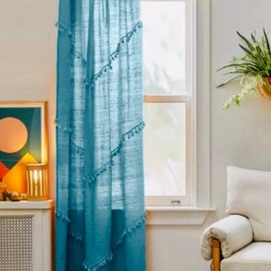 May include: A teal curtain hangs in front of a window, with a gold rod at the top. The curtain has a textured appearance and is adorned with small, white tassels arranged in diagonal rows. The curtain is in a room with a white wall and a patterned rug.