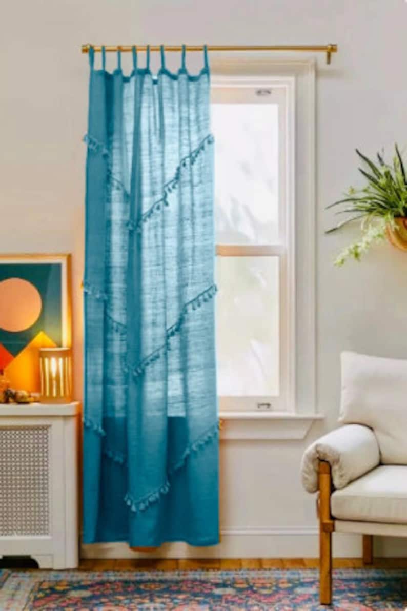 May include: A teal curtain hangs in front of a window, with a gold rod at the top. The curtain has a textured appearance and is adorned with small, white tassels arranged in diagonal rows. The curtain is in a room with a white wall and a patterned rug.