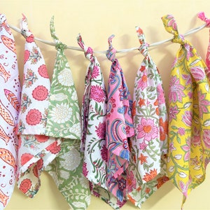 May include: A collection of colorful, patterned fabric napkins hanging from a white string. The napkins feature floral, paisley, and fish designs in shades of pink, green, yellow, and red. The napkins are knotted at the top.